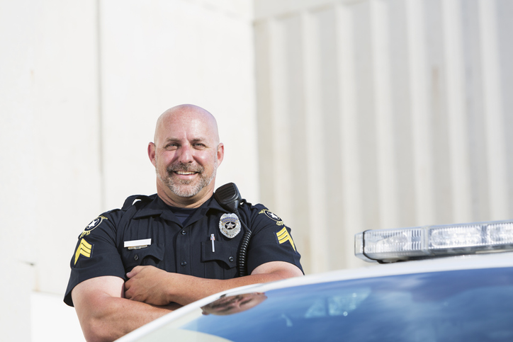 A police officer in uniform stands with arms crossed and smiles next to a police car, with a building and bright daylight in the background.
