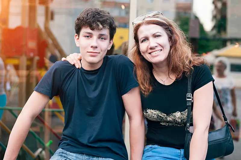 A smiling woman with long curly hair sits next to a teenage boy; her arm is around his shoulder. Both wear black t-shirts and are outdoors, with blurred buildings and people in the background.
