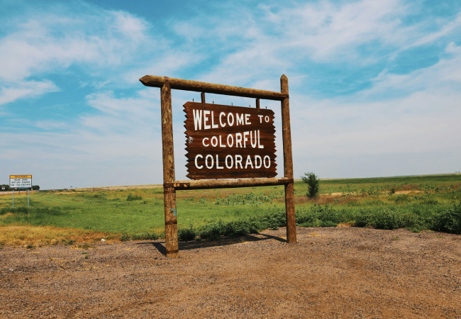A rustic wooden sign reads Welcome to Colorful Colorado, standing by a roadside with green fields and blue sky in the background.