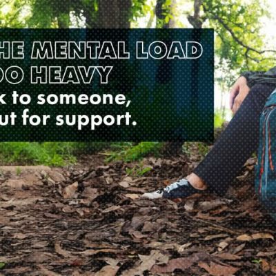A person sits in a forest clearing beside a large backpack, with text encouraging people to pause, talk to someone, or seek support when the mental load feels too heavy.