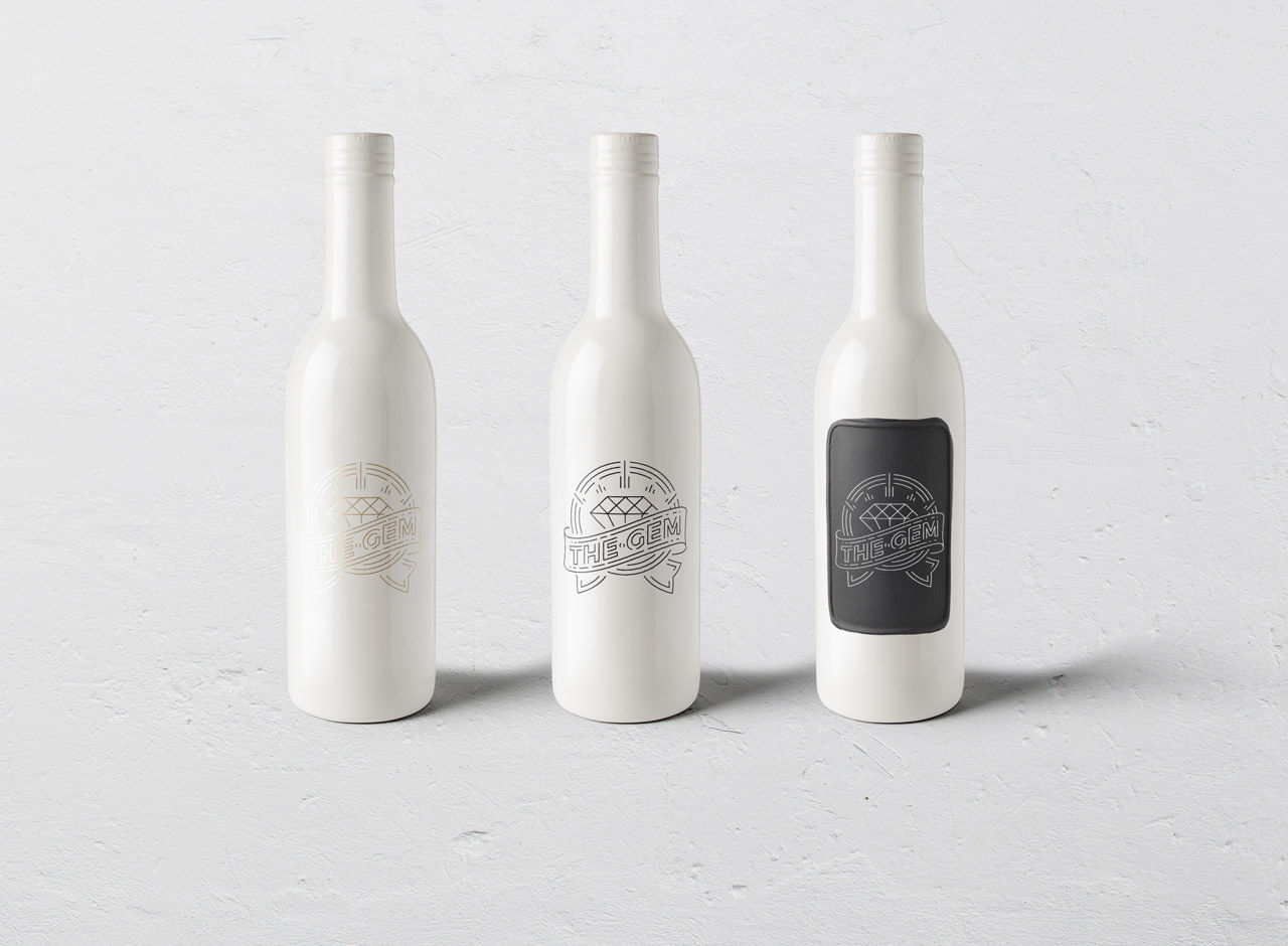 Three white wine bottles with cork tops are displayed side by side against a light gray background. Each bottle features a logo; the middle bottle has a clearer logo, and the right bottle has a black label with white design.
