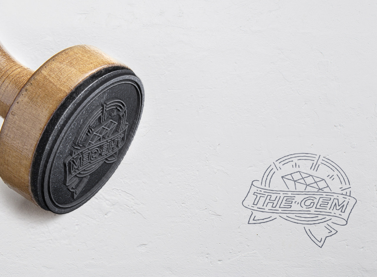 A rubber stamp with a wooden handle lies on its side, next to its stamped imprint on white paper. The stamp design features a diamond and the text “THE GEM” inside a ribbon banner.