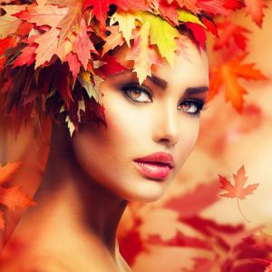 A woman with striking makeup wears a vibrant crown made of autumn maple leaves. Warm red, orange, and yellow tones create an autumnal, artistic atmosphere with falling leaves in the background.