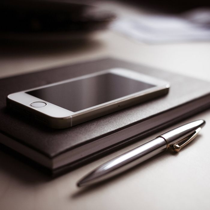 A silver pen rests beside a closed black notebook with a white smartphone placed on top, all on a light-colored surface.