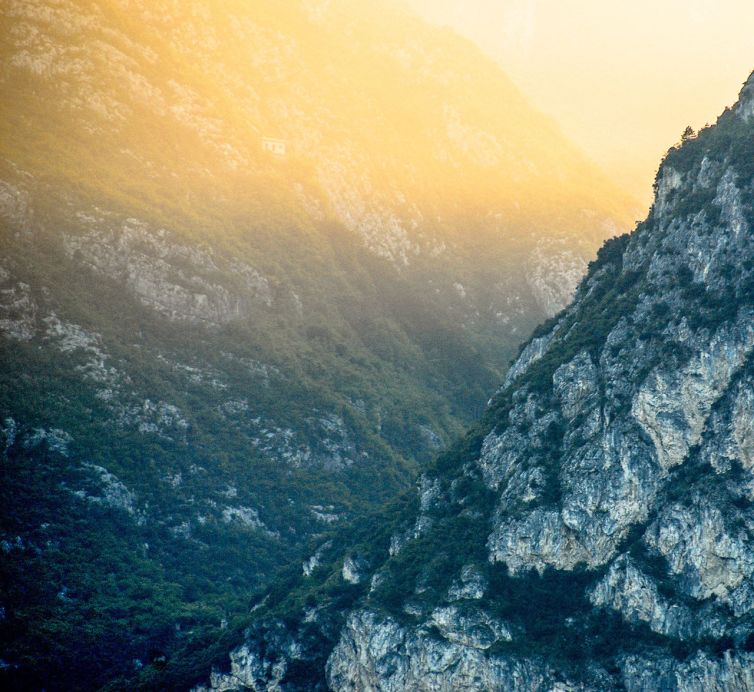 Steep rocky mountains covered with patches of greenery, bathed in golden sunlight. A small white house is visible in the distance, nestled high on the misty slope.