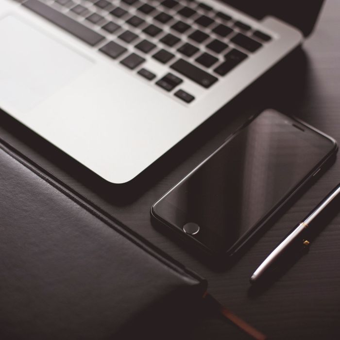 A close-up of a laptop keyboard, a smartphone, a closed notebook, and a pen arranged neatly on a dark desk.