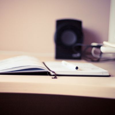 A pen rests on an open notebook on a wooden desk, with a blurred speaker and some cables in the background. The scene appears softly lit and calm, suggesting a workspace or study area.
