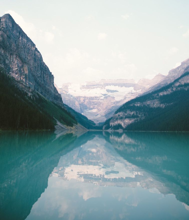 A tranquil turquoise lake reflects surrounding pine-covered mountains and distant snow-capped peaks under a hazy sky. The scene is serene and untouched, with soft, diffused lighting.