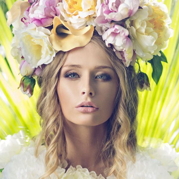 A woman with long blonde hair wears a large floral crown of white, yellow, and pink flowers. She poses in front of bright green leaves, gazing directly at the camera with a calm expression.