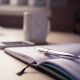 A close-up of an open notebook with a pen resting on its pages, a blurred mug, and a smartphone in the background on a light-colored desk.