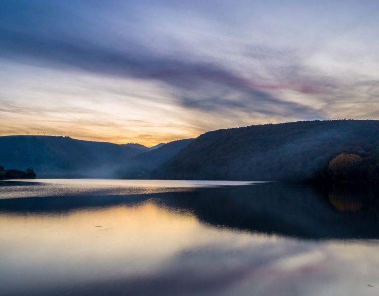 A calm lake reflects the soft colors of dusk, with gentle hills and forests silhouetted against a cloudy, pastel sky. The tranquil water mirrors the scenic landscape and fading light.