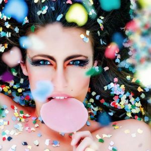 A woman lies surrounded by colorful confetti, holding a pink lollipop near her mouth. Her makeup is dramatic, with bold eye shadow and long eyelashes, creating a striking and festive look.