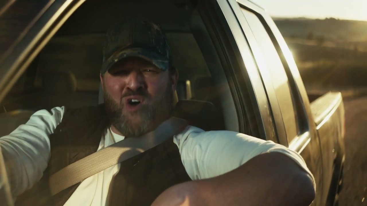 A bearded man drives a pickup truck with the driver's side window rolled down and his arm rested on the windowsill, while mouthing words.