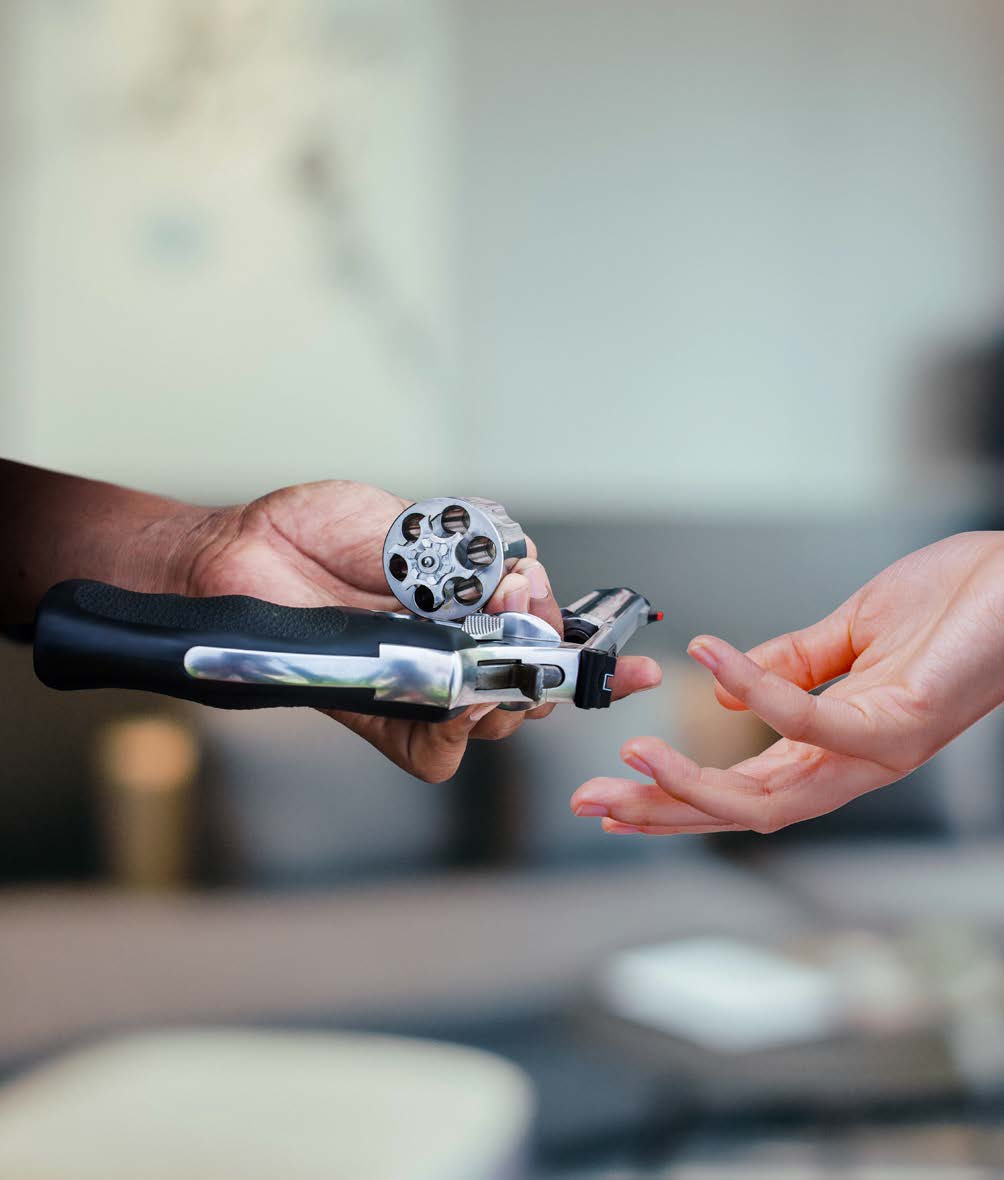 Una persona entrega un revólver con el cilindro abierto y vacío a otra, cuya mano se extiende para recibirlo. El fondo está difuminado, centrando la atención en las manos y el revólver.