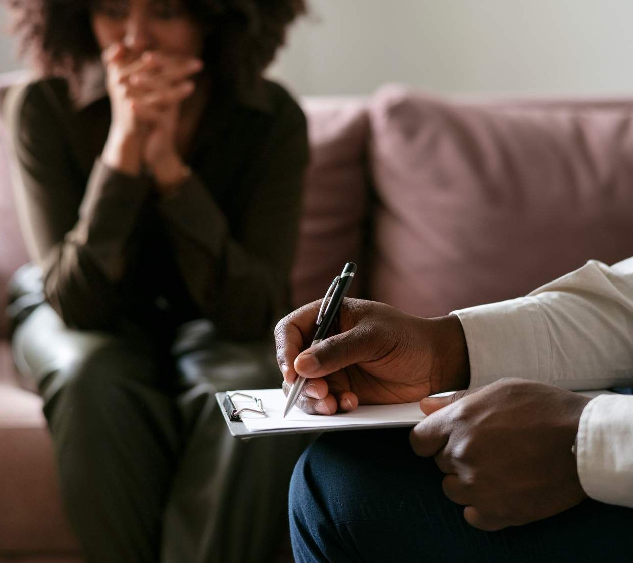Close-up of a person taking notes on a clipboard during a counseling session. In the background, a woman sits on a couch with her hands clasped near her mouth, appearing deep in thought or concerned.