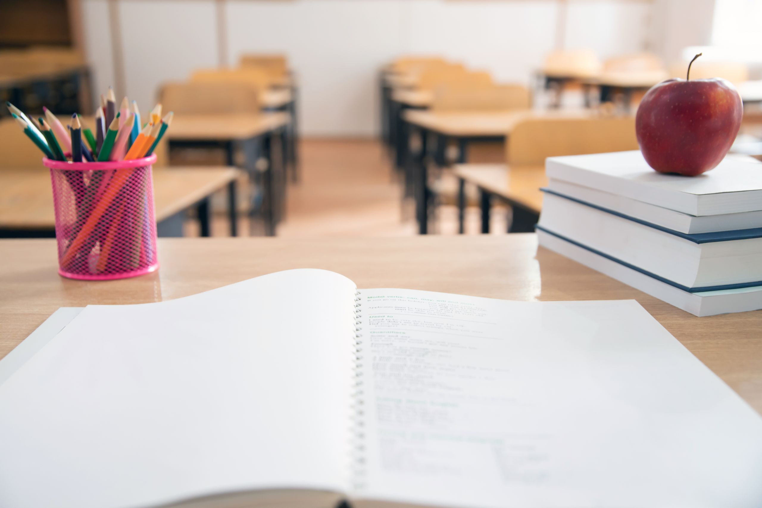 An open notebook sits on a classroom desk next to a cup of colored pencils and a stack of books topped with a red apple, with blurred rows of desks in the background.