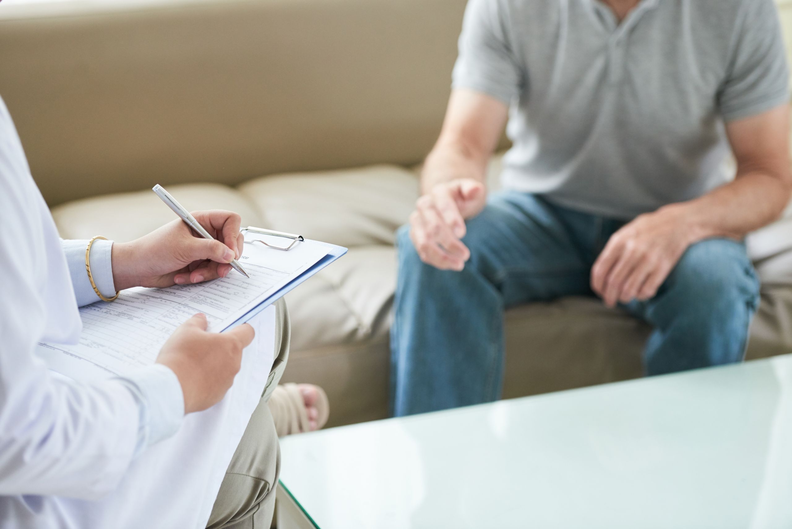 A healthcare provider in a white coat writes on a clipboard while a patient sits across from them.